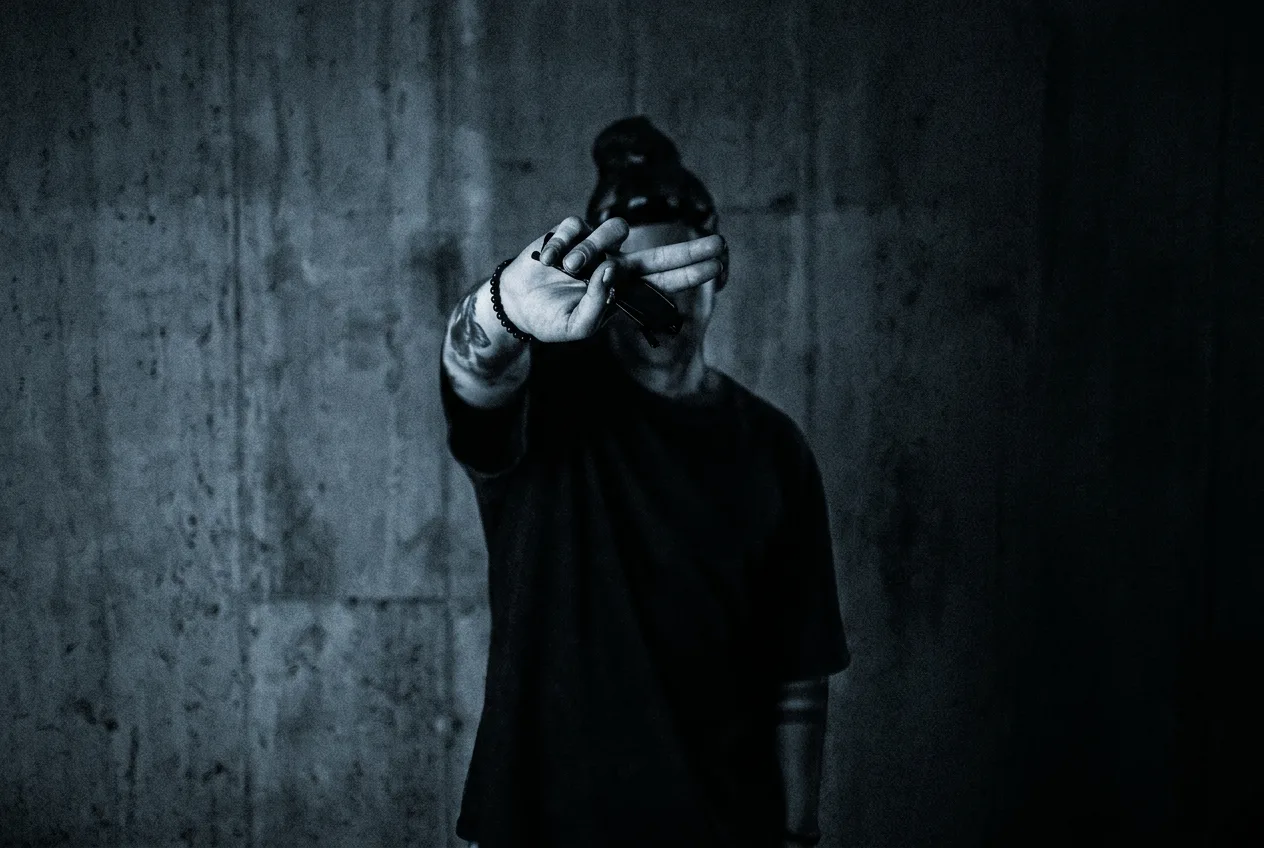 DAY SHO shielding his face with an outstretched hand in front of a concrete wall